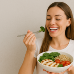 Junge Frau mit hellem Shirt lächelt und isst Brokkoli aus einer Gabel, während sie eine Schale mit Spinat, Hähnchen, Quinoa und Tomaten in den Händen hält